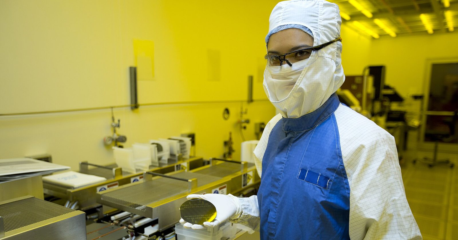 Technik w białym kombinezonie i masce trzyma okrągłą płytkę krzemową w żółto oświetlonym laboratorium czystym