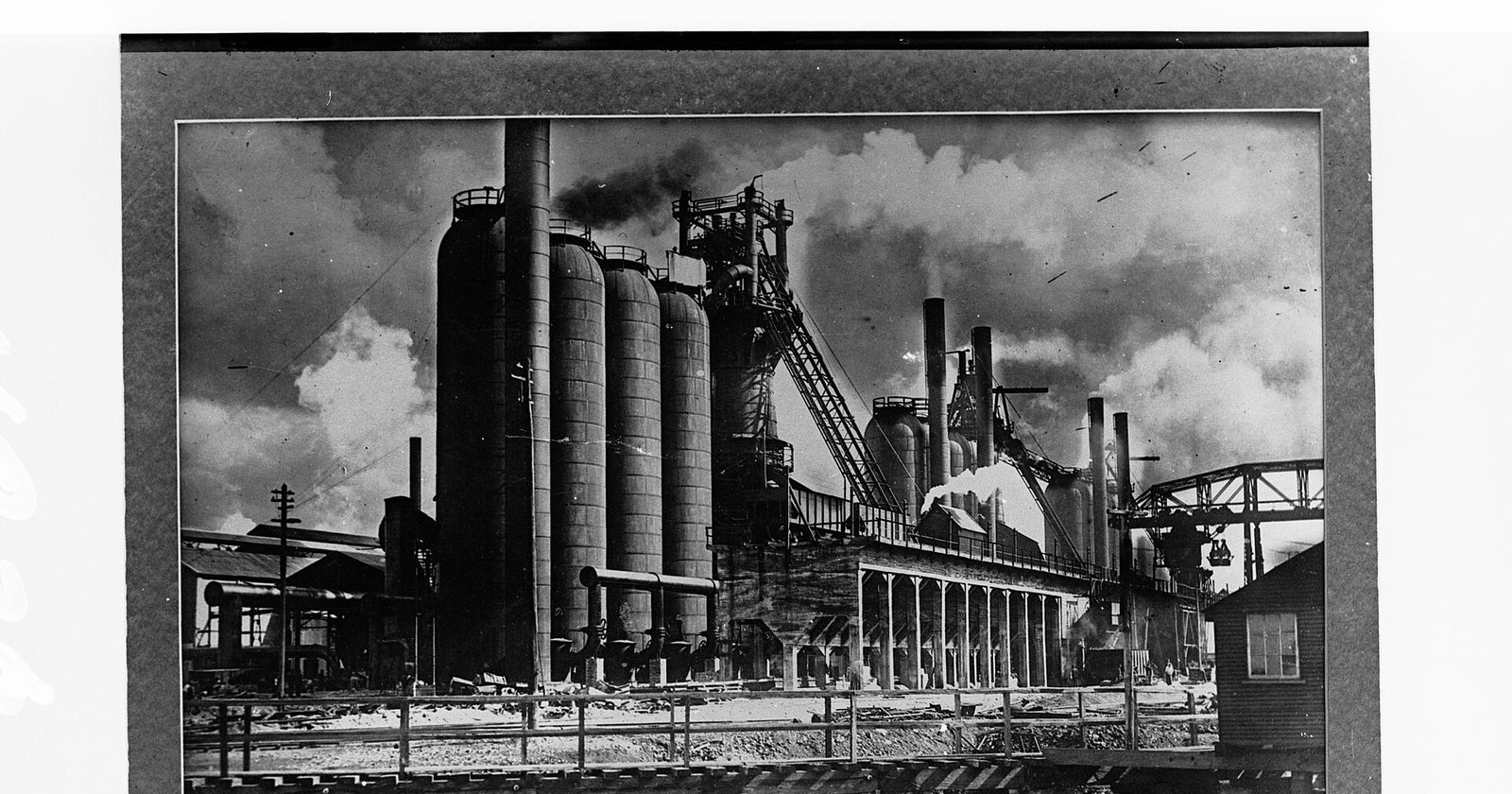 Archiwalna czarno-biała fotografia trzech wielkich pieców hutniczych z kominami, estakadą i dymem nad zakładem